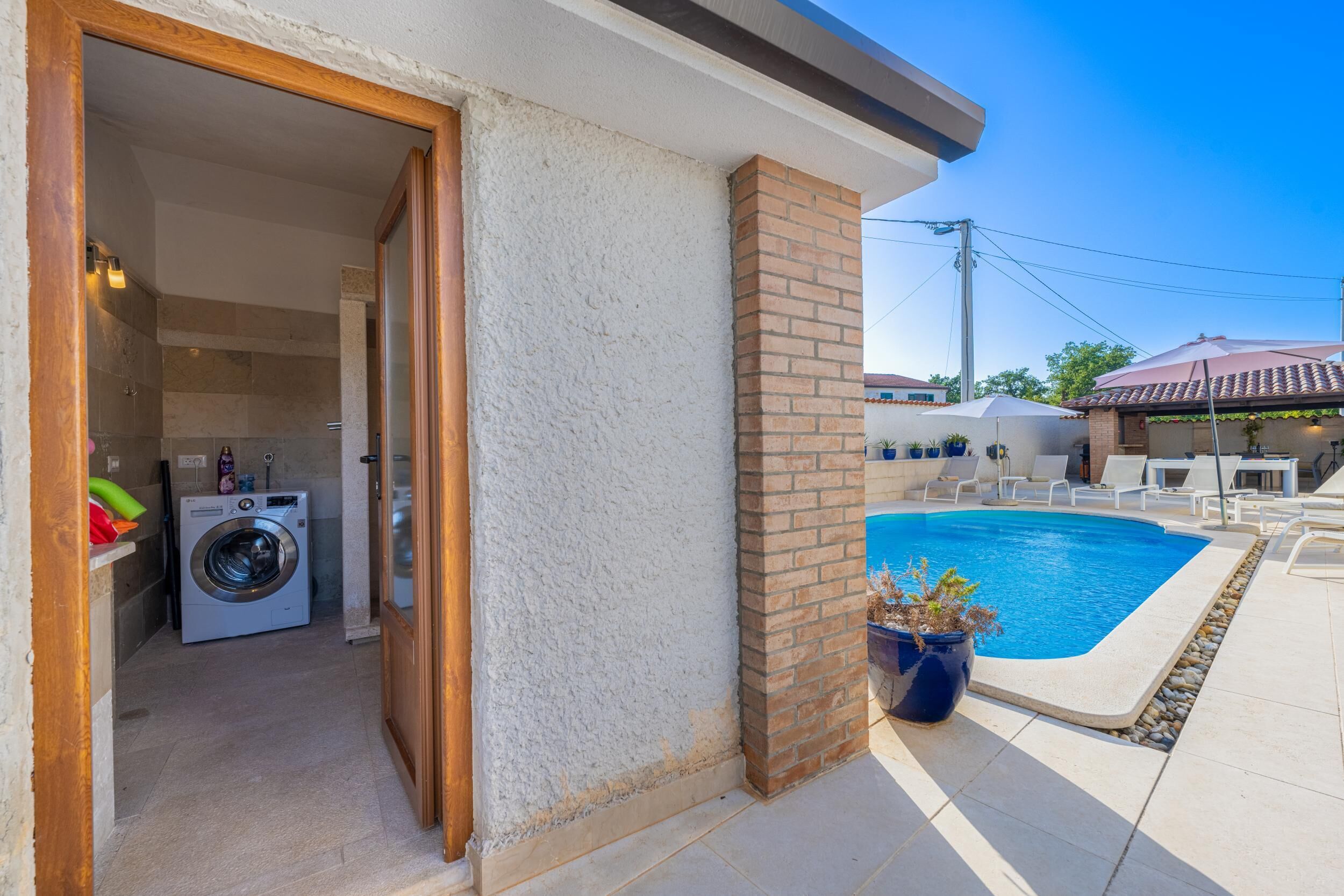 A poolside area with a clear view of a swimming pool, a brick column, and a washing machine in the background.