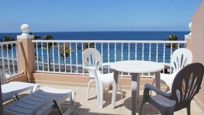A balcony with white plastic chairs and a table, overlooking a beach with palm trees and the ocean.