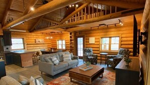 A spacious log cabin interior with a high ceiling, a dining area, a living room with a sofa and armchairs, and a kitchen area. - Aspen Bear Paws | Charming Retreat with Hot Tub & Loft (Alpine)