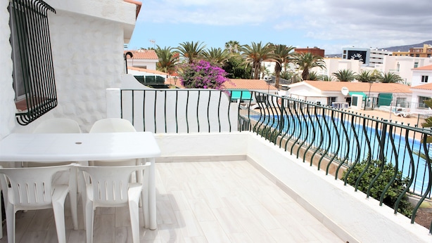 A balcony with a white table and chairs, a pool, and a view of buildings and palm trees.