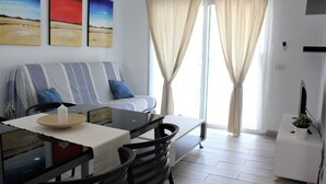 A modern living room with a sofa, dining table, and chairs. There are three paintings on the wall, a window with curtains, and a TV on a stand.