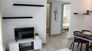 A modern living room with a flat-screen TV on a white console, a small potted plant, and a dining area with a table and chairs.