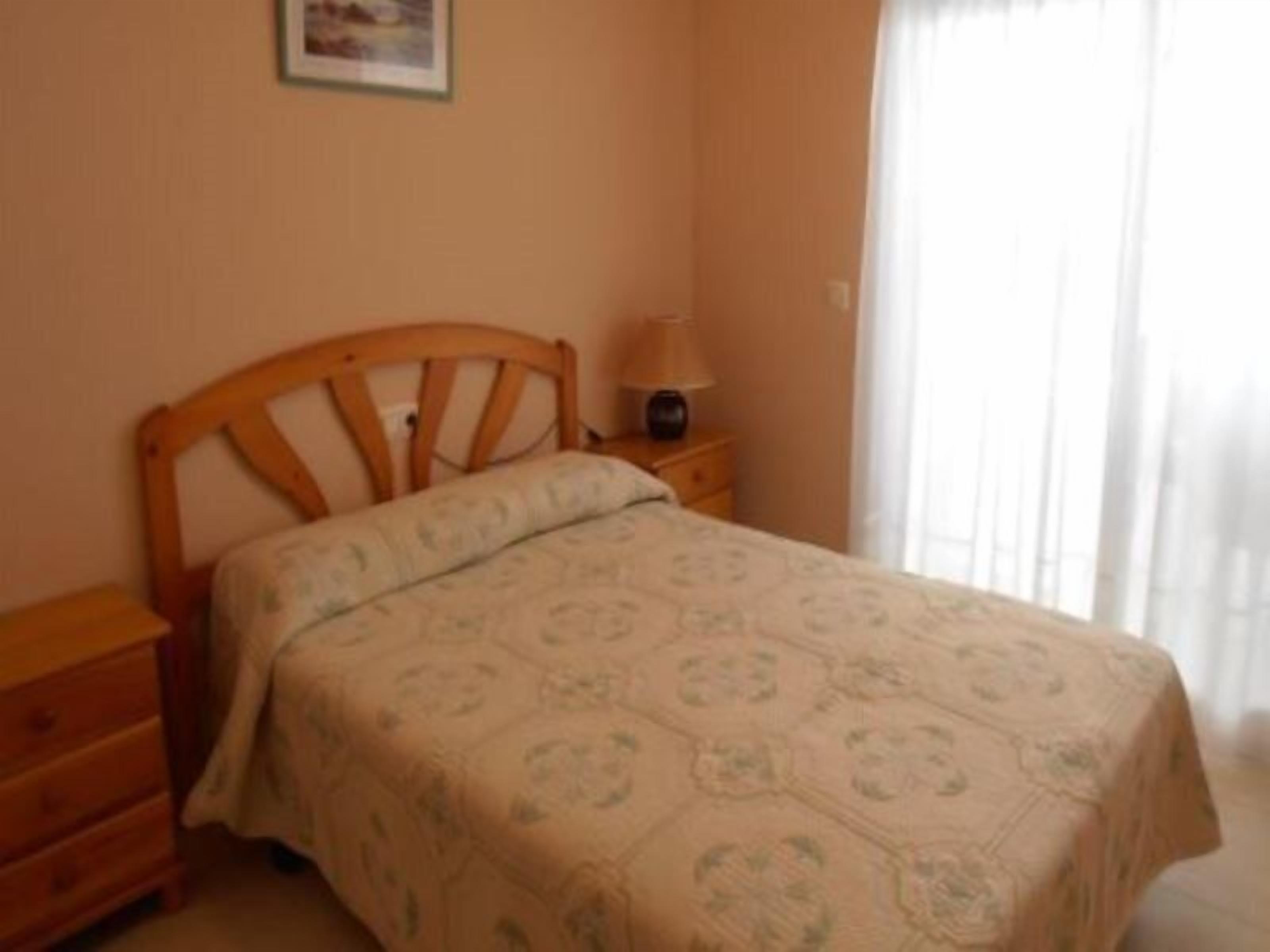 A bedroom with a wooden bed, a bedside table, a lamp, and a window with curtains.