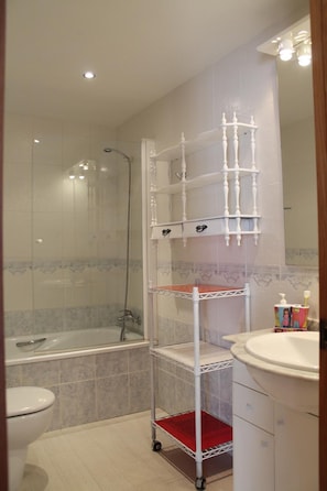 A bathroom with a shower, a toilet, a sink, and a white metal shelf unit. - Argenta Caleta Apartaments (Peniscola)