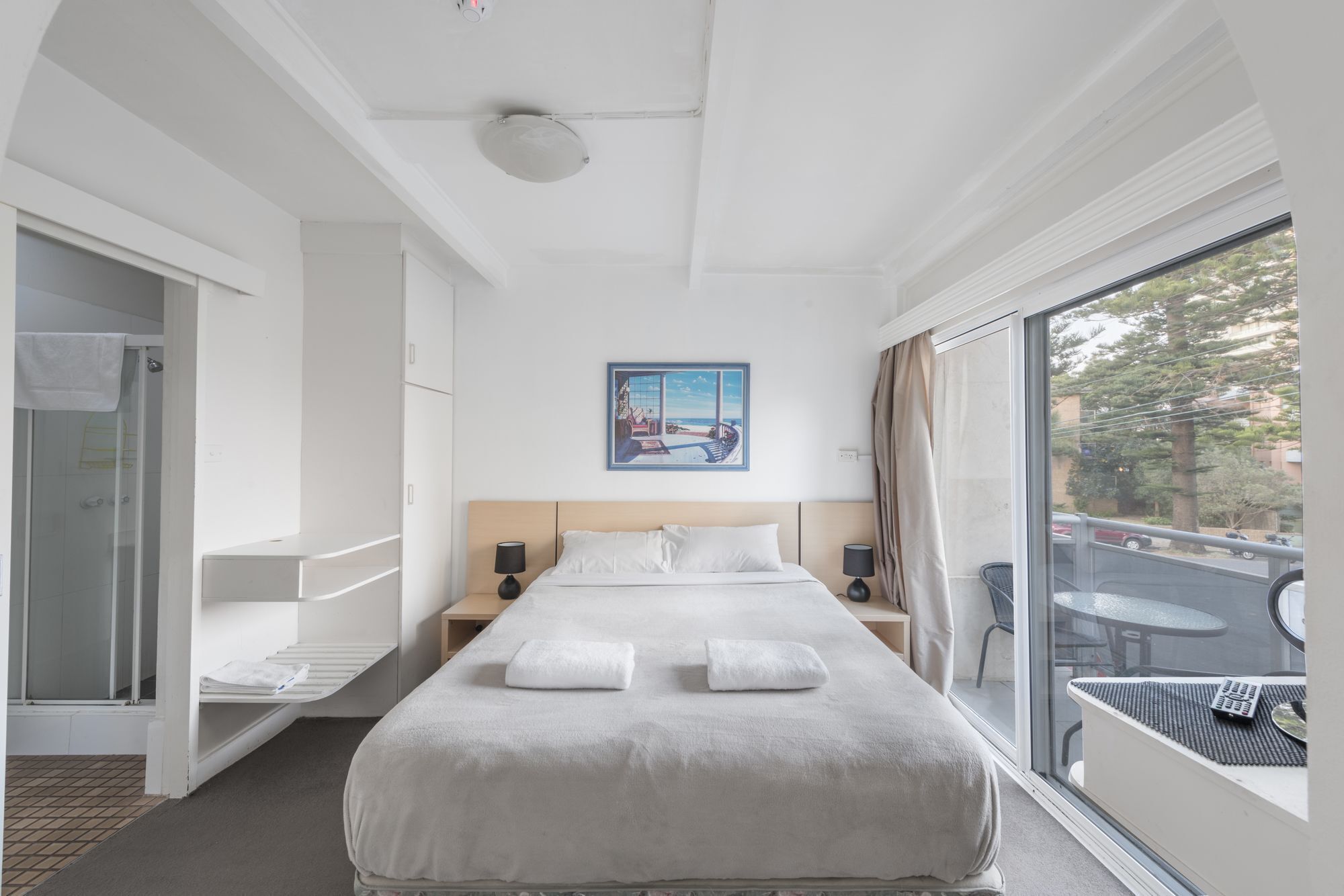 A hotel room with a large bed, a painting above the bed, a view of the outside through a large window, and a shower area to the left.