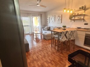 A modern living room with a sofa, dining table, and chairs. There is a kitchen area with a stove and shelves. - Mareverde spacious 1 bedroom apartment (Costa Adeje)