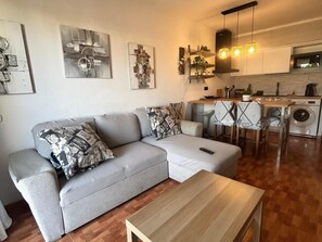 A modern living room with a grey sectional sofa, a wooden coffee table, and a kitchen area with a washing machine.