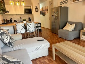 A modern living room with a sofa, armchair, and coffee table. There is a kitchen area with a microwave, sink, and cabinets. A refrigerator is visible in the background.