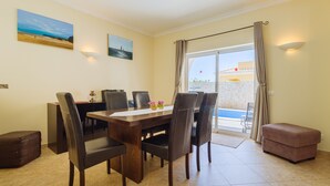 A dining area with a wooden table and chairs, a small ottoman, and two paintings on the wall. - Poolvilla Casa Vista Mar (Carvoeiro)
