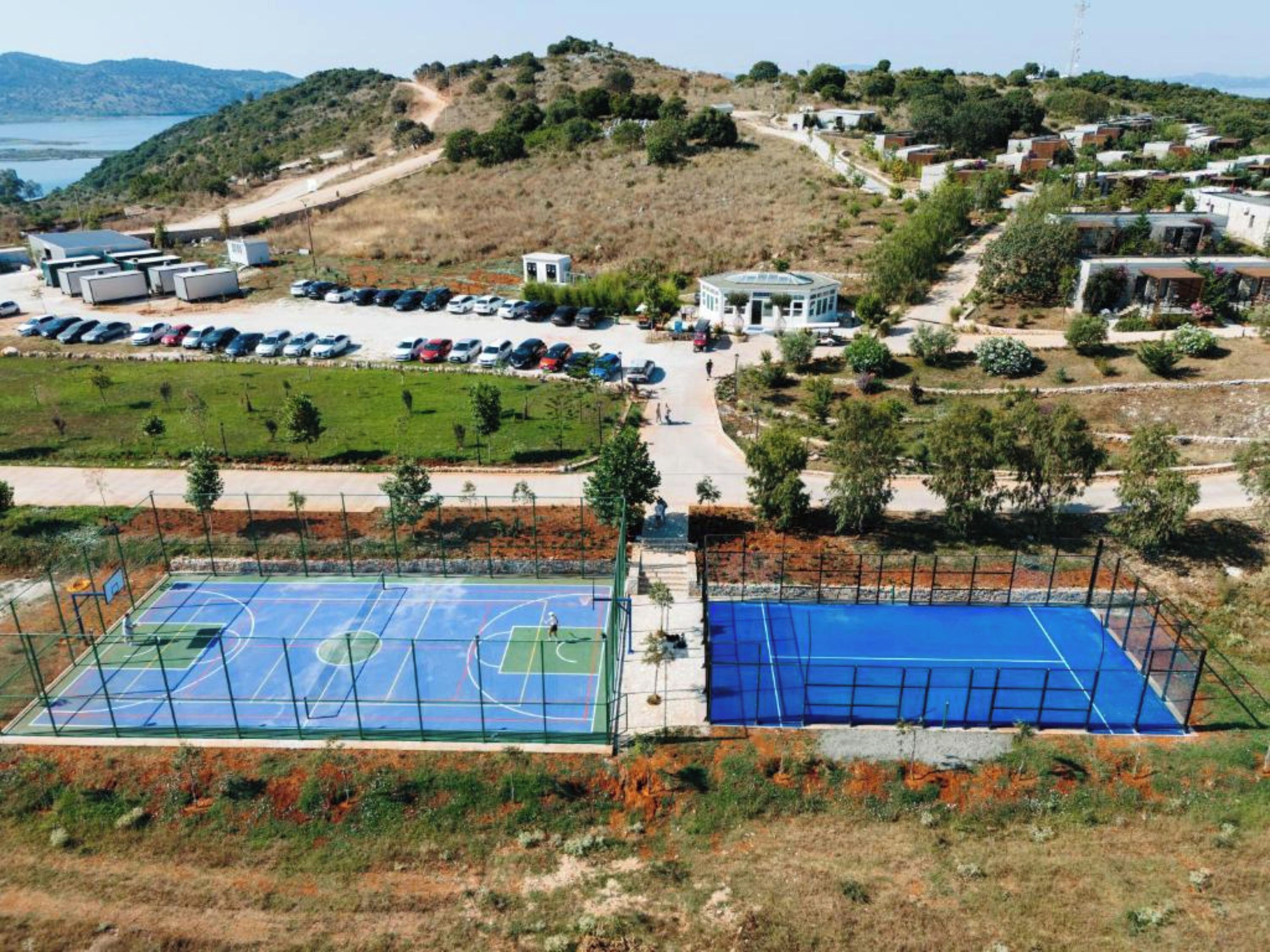 A basketball court with a blue surface, surrounded by a fence, and a parking lot with several cars.