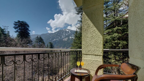 A balcony with a view of mountains, a chair, and a small table with two glasses of drink. - Regenta Place Green Leaf Manali- A Centrally Heated Resort (Manali)