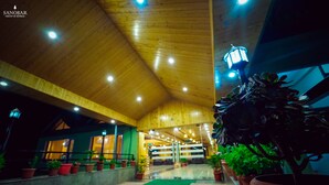 A spacious indoor area with a high wooden ceiling and multiple ceiling lights. There are potted plants along the walls and a green mat on the floor. - Regenta Place Green Leaf Manali- A Centrally Heated Resort (Manali)