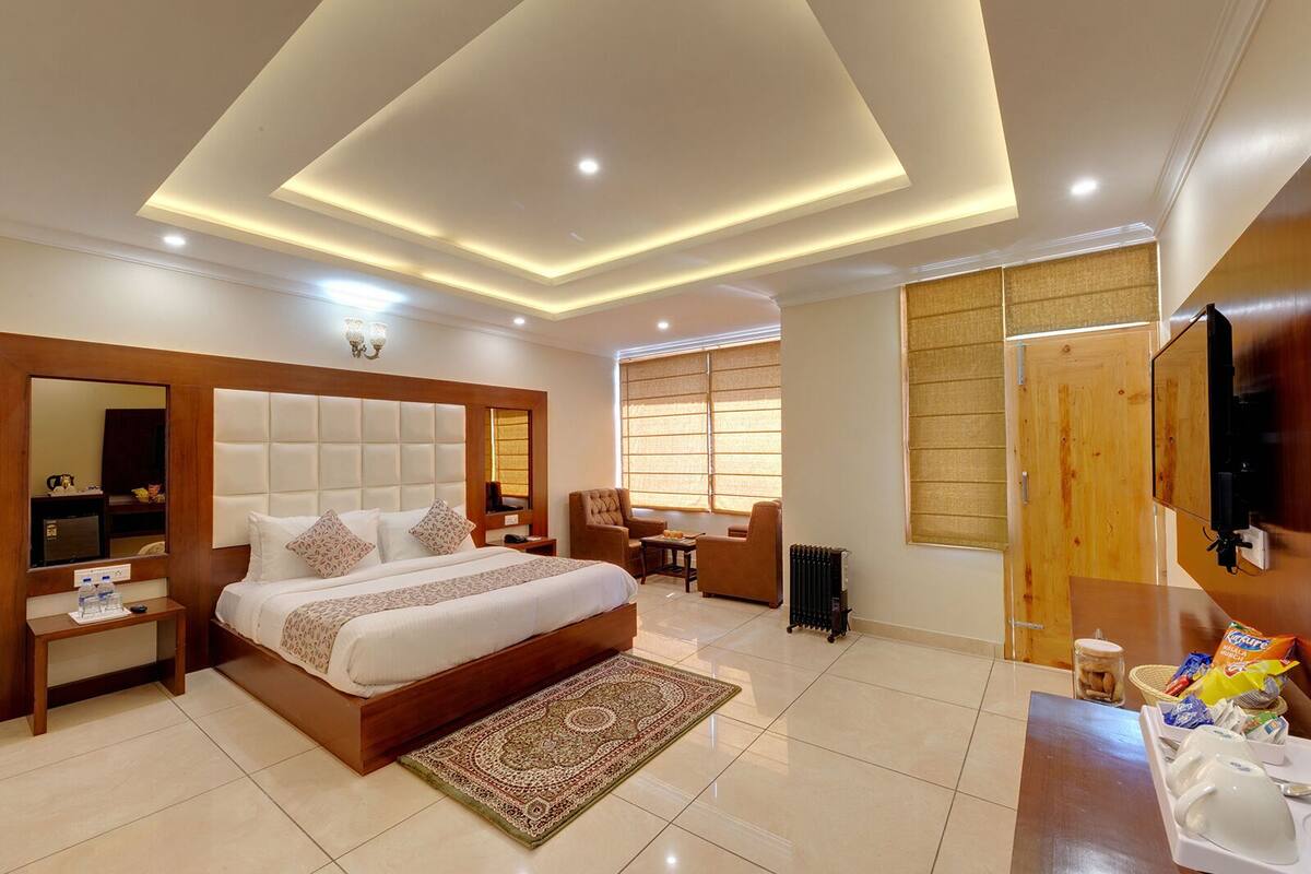 A modern hotel room with a large bed, a sitting area with a chair and table, a television, and a large window with blinds.