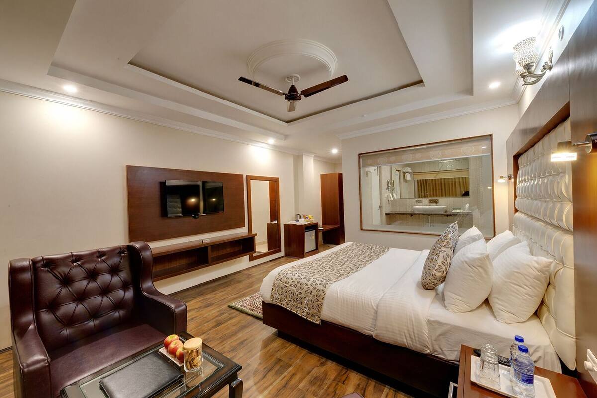 A modern hotel room with a large bed, a leather armchair, a TV, a mirror, and a ceiling fan.