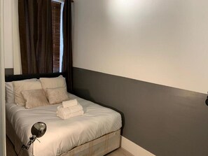 A neatly made bed with pillows and folded towels, a bedside lamp, and a window with curtains. - Portobello Stays by Stay Guru (London)