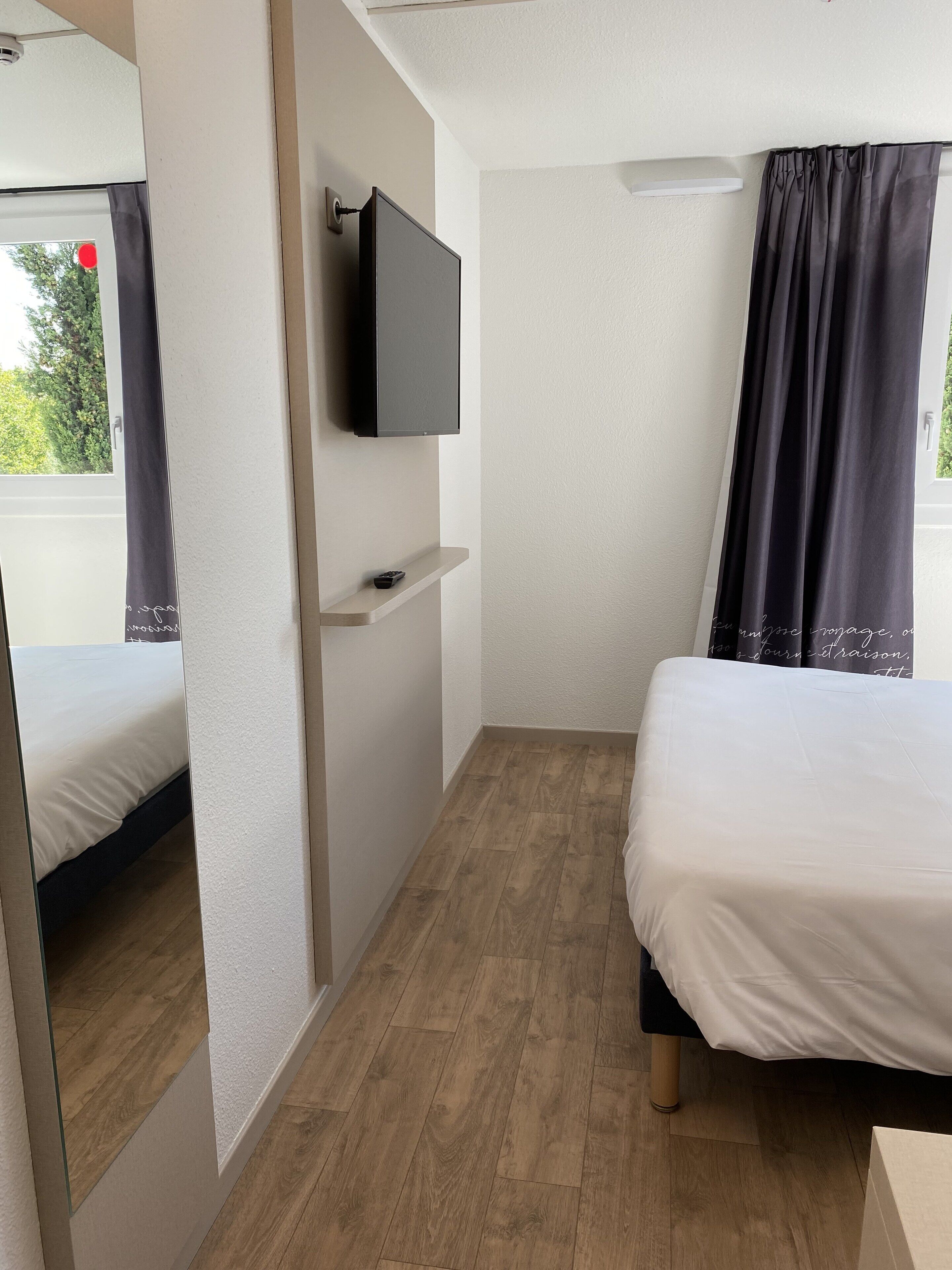 A bedroom with a bed, a TV mounted on the wall, a shelf, and a window with curtains.
