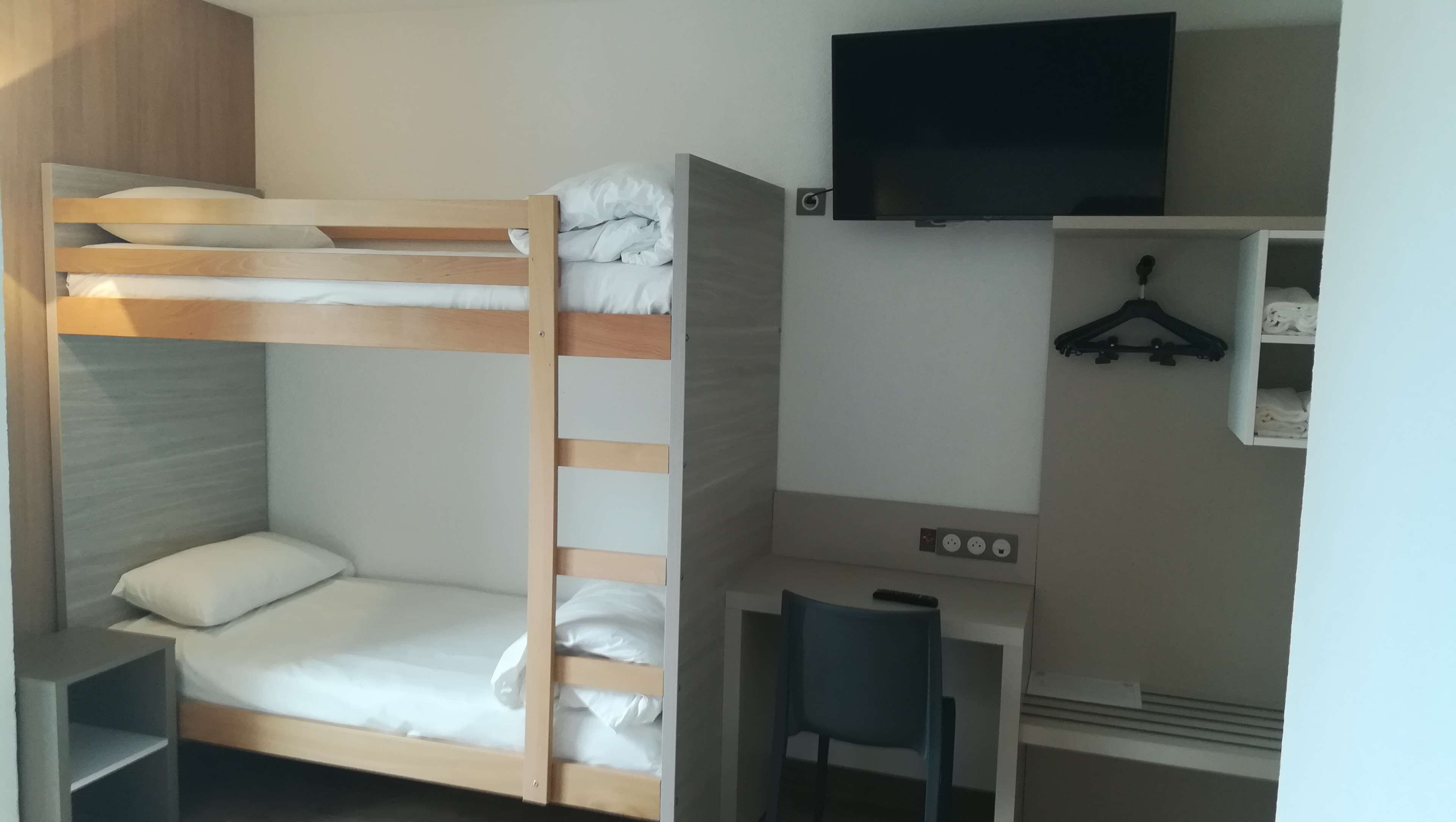 Una cama litera con escritorio y silla, un televisor montado en la pared y un armario con perchas.
