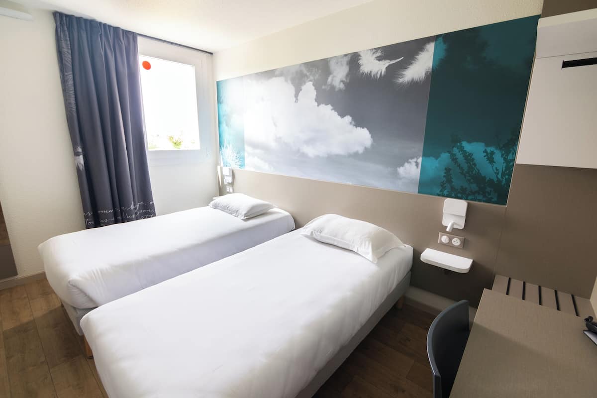 A hotel room with two single beds, a desk, a chair, a window with curtains, a wall-mounted phone, and a wall art depicting clouds.