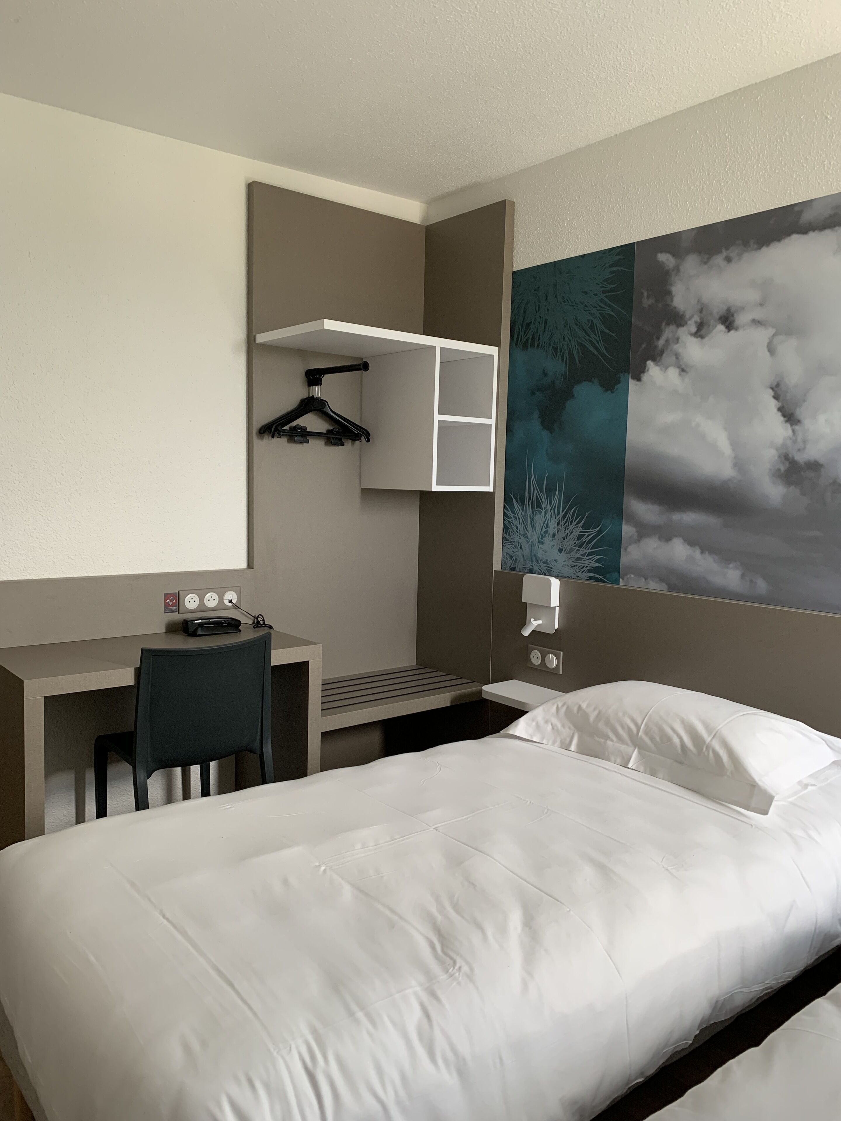 Une chambre d’hôtel équipée d’un lit, d’un bureau, d’une chaise et d’une étagère fixée au mur, avec un cintre noir.