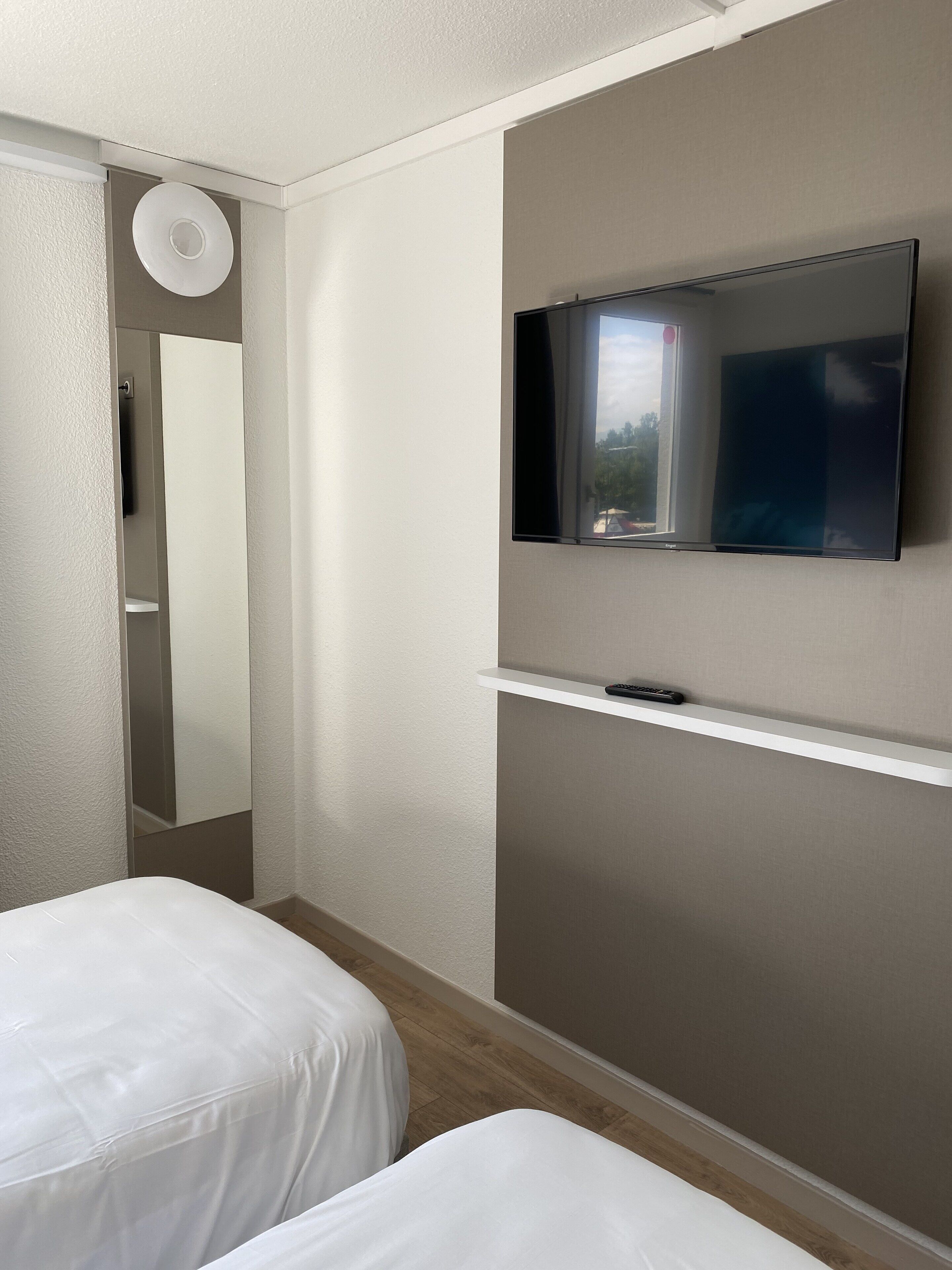 Habitación de hotel con televisor de pantalla plana montado en la pared, un estante pequeño debajo y dos camas con ropa blanca.