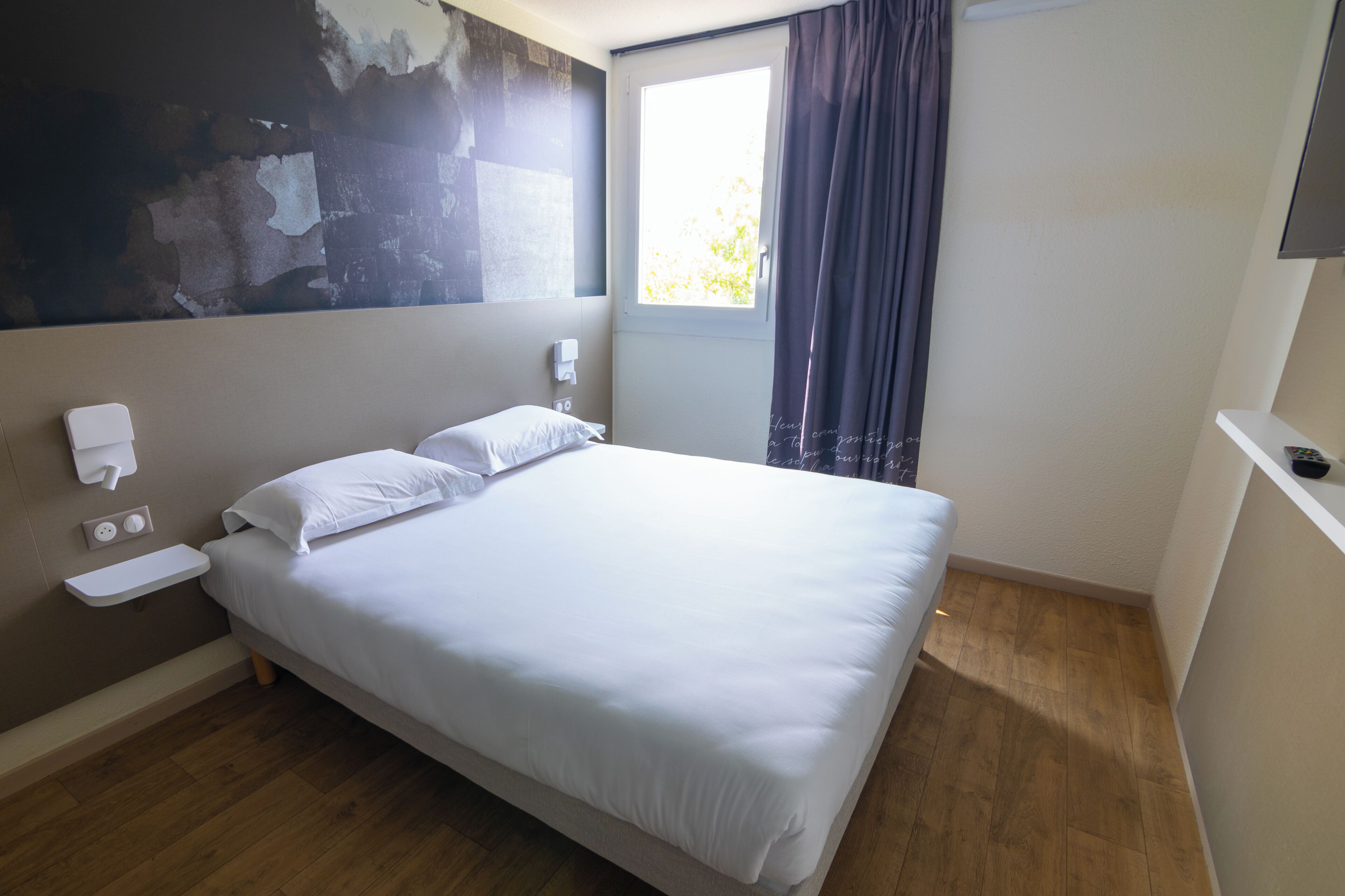 Una habitación de hotel con cama, mesita de noche, teléfono en la pared y ventana con cortinas.