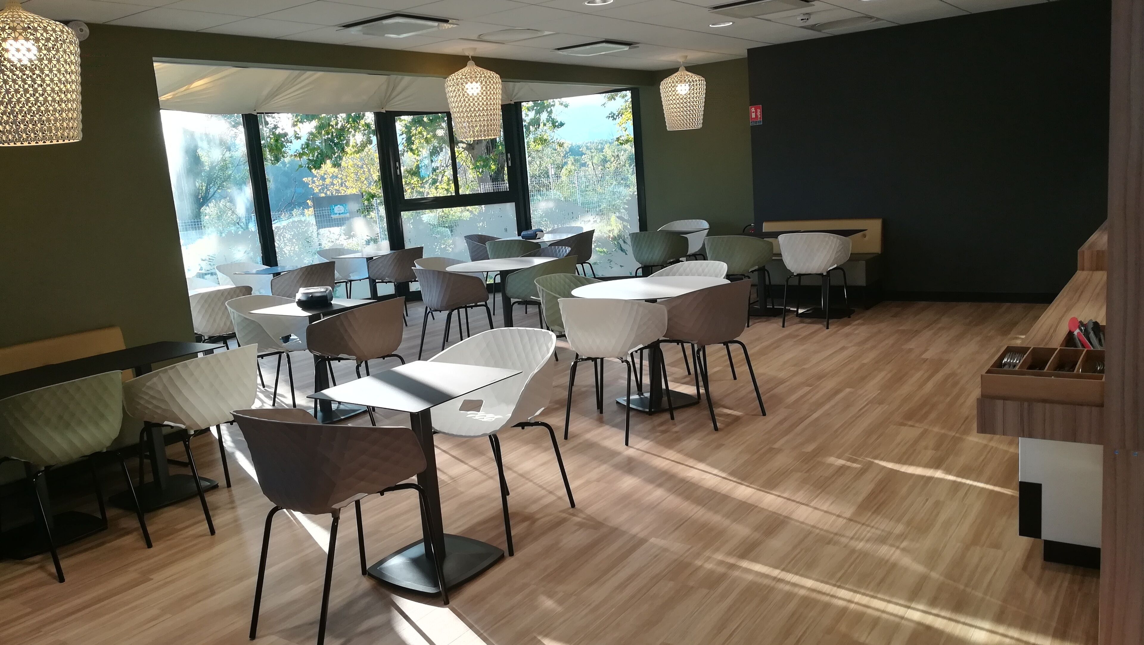 Un restaurant doté de nombreuses tables et chaises, de grandes fenêtres offrant une vue sur les arbres et de luminaires suspendus.