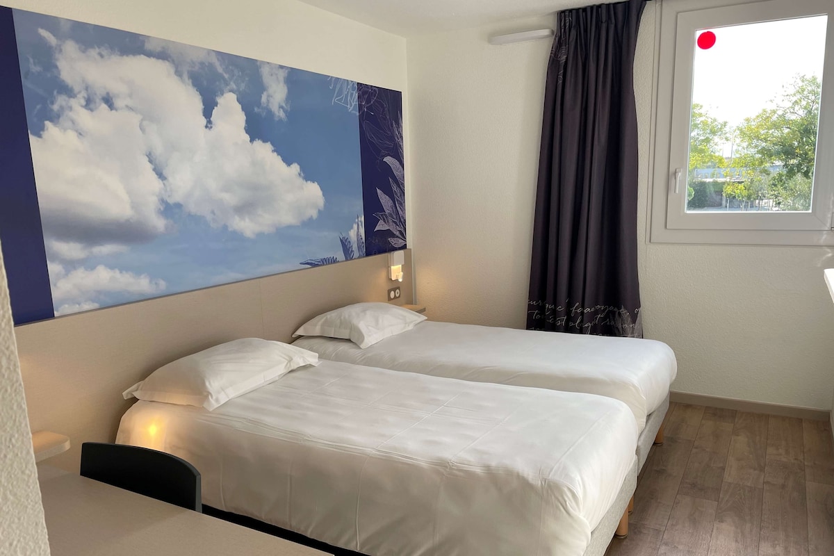 Une chambre d’hôtel avec deux lits, un bureau, une grande fresque murale et une fenêtre avec des rideaux.
