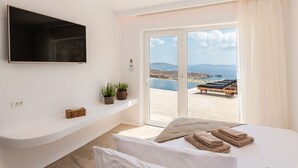 A modern bedroom with a bed, a flat-screen TV, a small shelf with plants, and a view of the sea through a sliding glass door. - Breathtaking 5 Bedroom Villa With Private Pool (Mykonos)