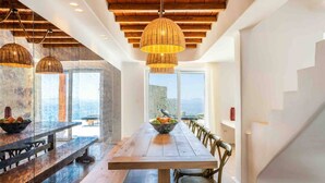 A modern dining area with a long wooden table, wicker pendant lights, and a view of the sea through large windows. - Breathtaking 5 Bedroom Villa With Private Pool (Mykonos)