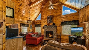 A log cabin interior with a fireplace, a red sofa, a television, and a stone fireplace surround. - Another Day In Bearadise- “Cozy Cabin Retreat in the Smokies – Comfort & Fun!!! (Pigeon Forge)