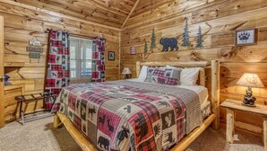 A cabin interior with a wooden bed, a window with plaid curtains, and wall decorations including a bear and pine tree. - Another Day In Bearadise- “Cozy Cabin Retreat in the Smokies – Comfort & Fun!!! (Pigeon Forge)