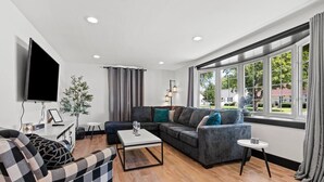 A modern living room with a sofa, a coffee table, a TV, and a large window with curtains. - 5BR House by Lambeau with Game Garage (Green Bay)
