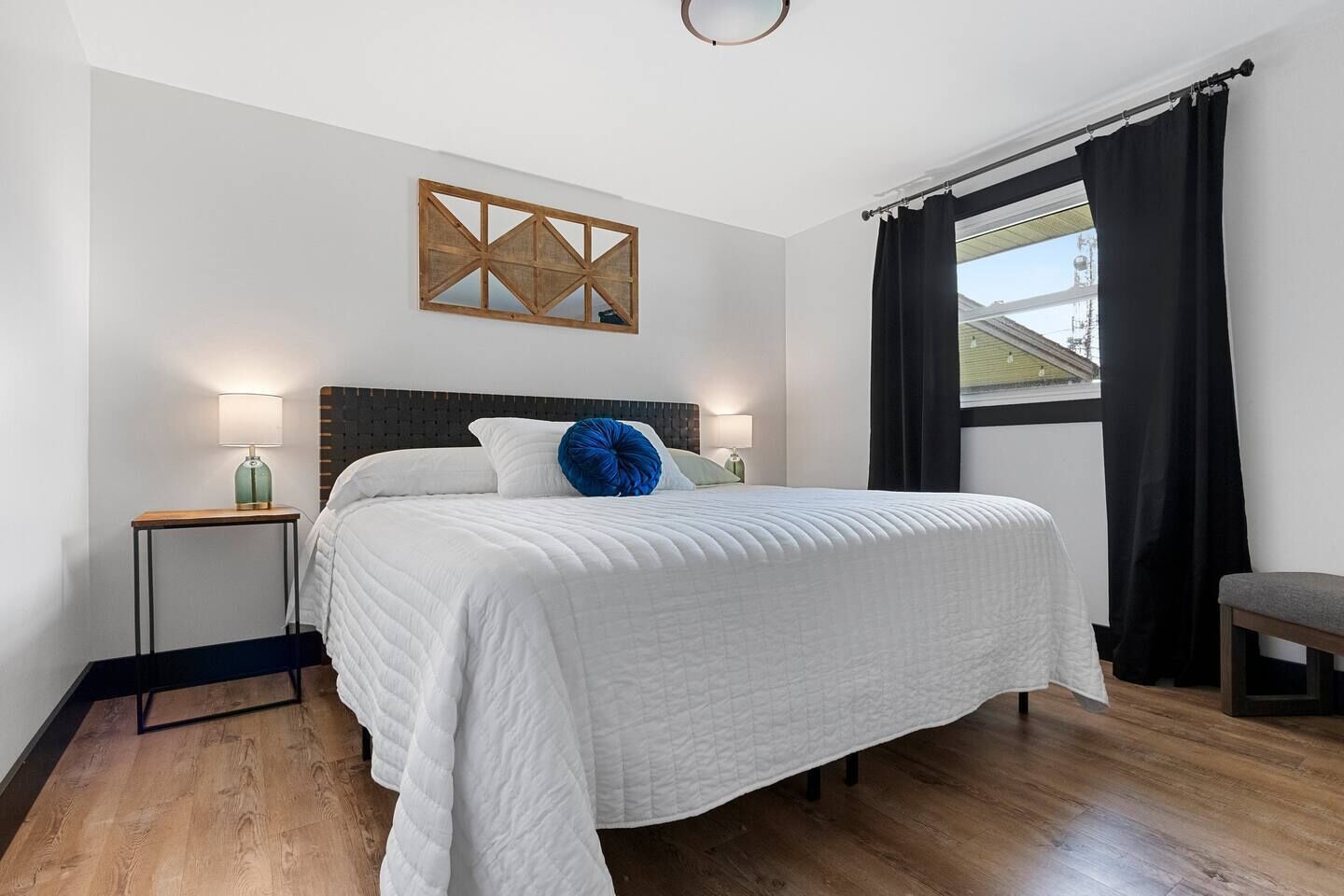 A bedroom with a large bed, a headboard, bedside tables, a window with curtains, and a wall-mounted artwork.