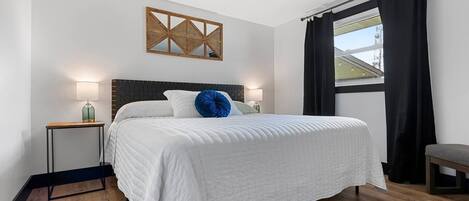 A bedroom with a large bed, a headboard, bedside tables, a window with curtains, and a wall-mounted artwork.