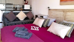 A hotel room with a bed, pillows, a bedside table, a small desk, and a kitchen area in the background. - Mareverde Sun apartment, pool view (Costa Adeje)