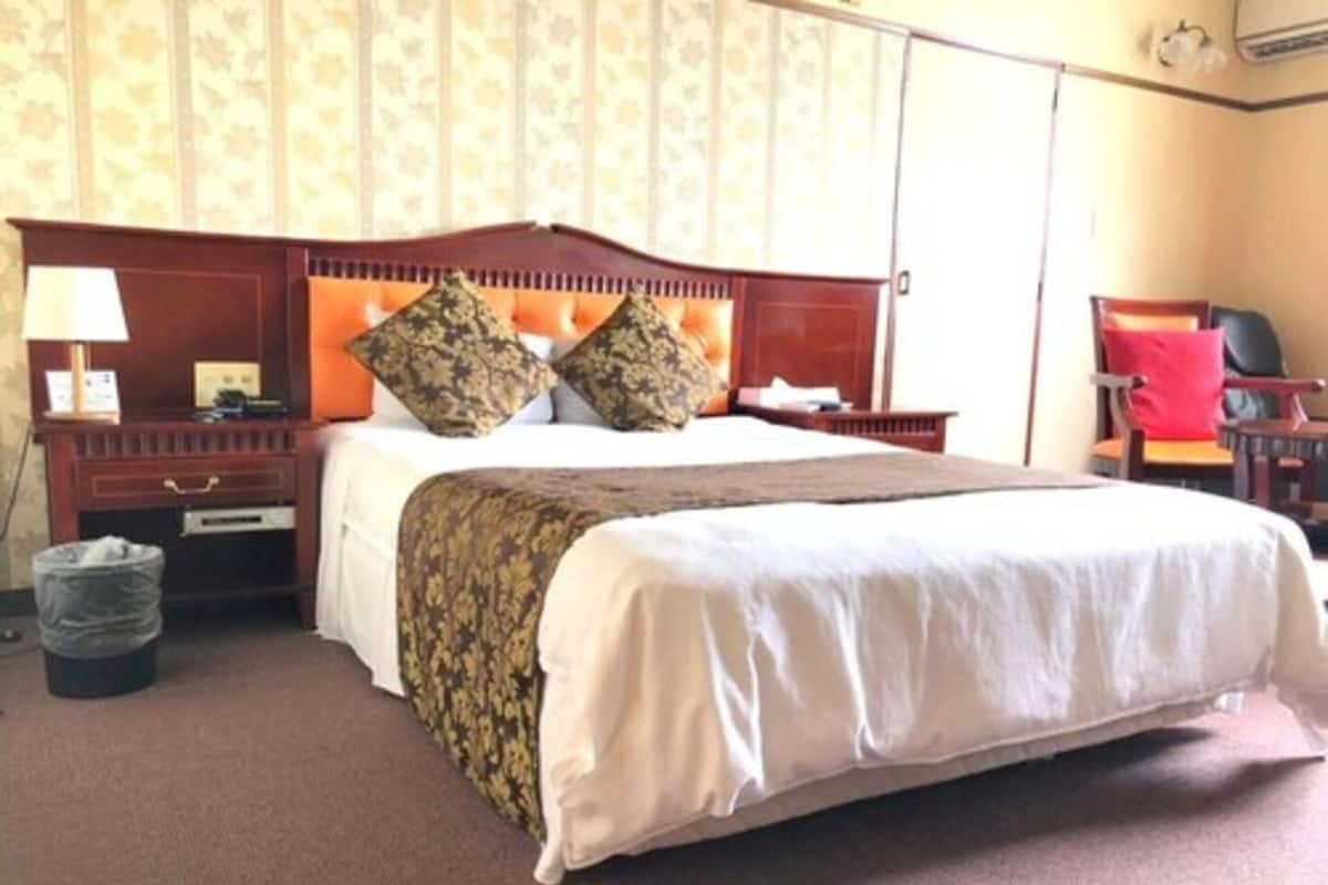 A hotel room with a large bed, a wooden headboard, a nightstand with a lamp, a chair, and a trash bin.