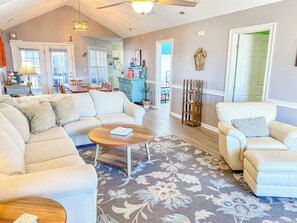 A living room with a sofa, armchair, and coffee table. There is a door leading to another room and a window with blinds.