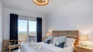 A hotel room with a large bed, a wooden headboard, a nightstand with a lamp, and a view of the beach through the window. - Front Row Penthouse Apartment on Carvajal Beach (Fuengirola)