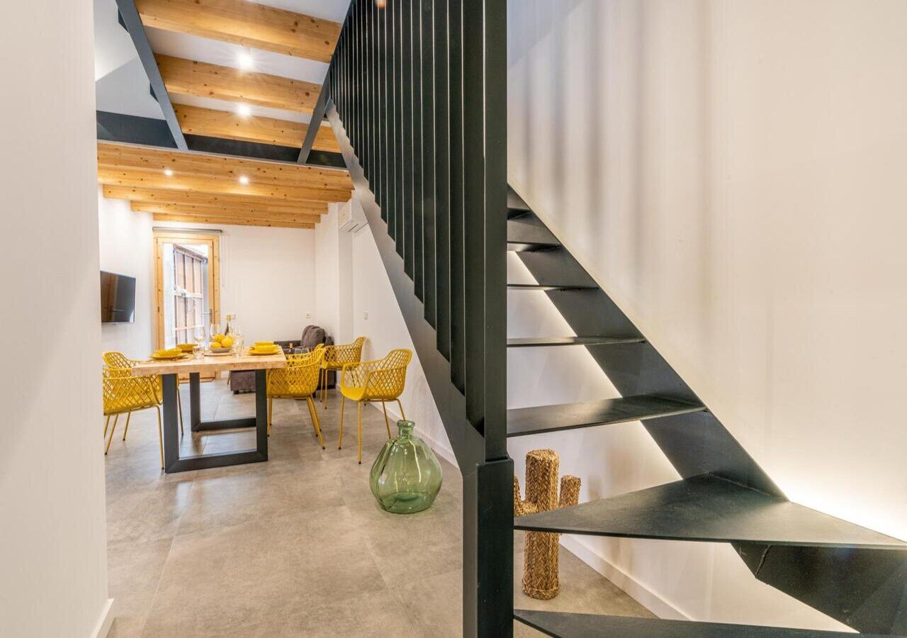 Un interior moderno con una escalera, una zona de comedor con sillas amarillas y un jarrón verde.
