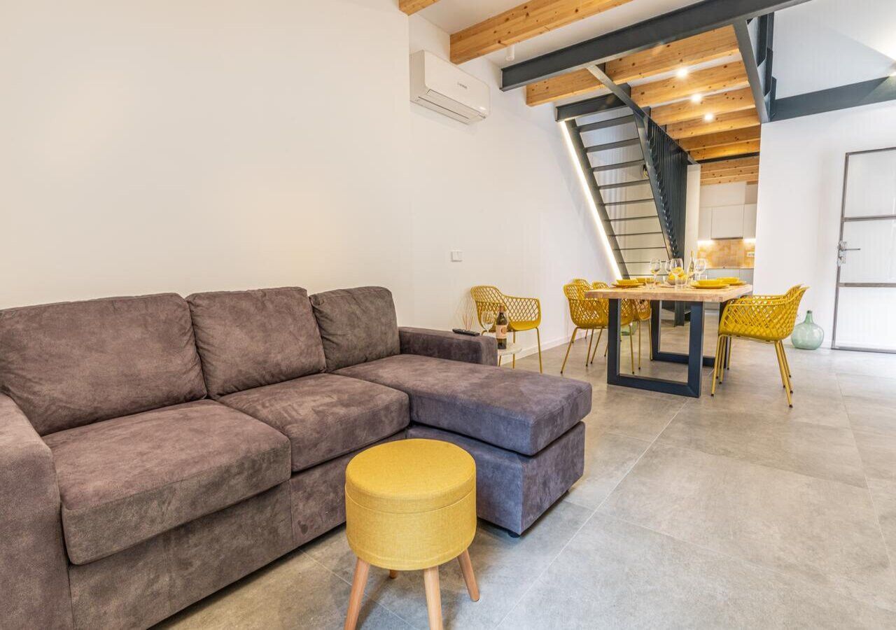 Ein modernes Wohnzimmer mit einem grauen Ecksofa, einem kleinen runden Hocker und einem Essbereich mit gelben Stühlen und einem Tisch.
