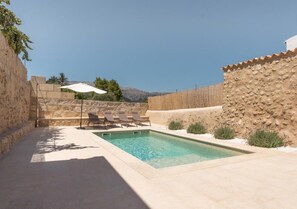 A rectangular pool with clear water, surrounded by a stone wall and a white fence. There are three lounge chairs and a white umbrella near the pool. - Beautiful Mallorcan house with 2 bedrooms, 1 bathroom, pool and barbecue (Campanet)