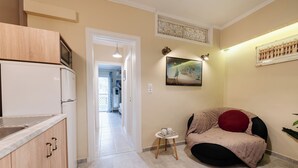A compact kitchen with a microwave, a sink, and wooden cabinets. A bean bag chair is placed in a corner, and a small table with a cup and saucer is nearby. A painting hangs on the wall, and a framed picture is also visible. - Palaiopolis Maison by Konnect, Mon Repos (Corfu)