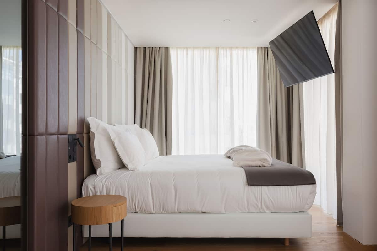 A modern bedroom with a large bed, a small round table, and a flat-screen TV mounted on the wall.