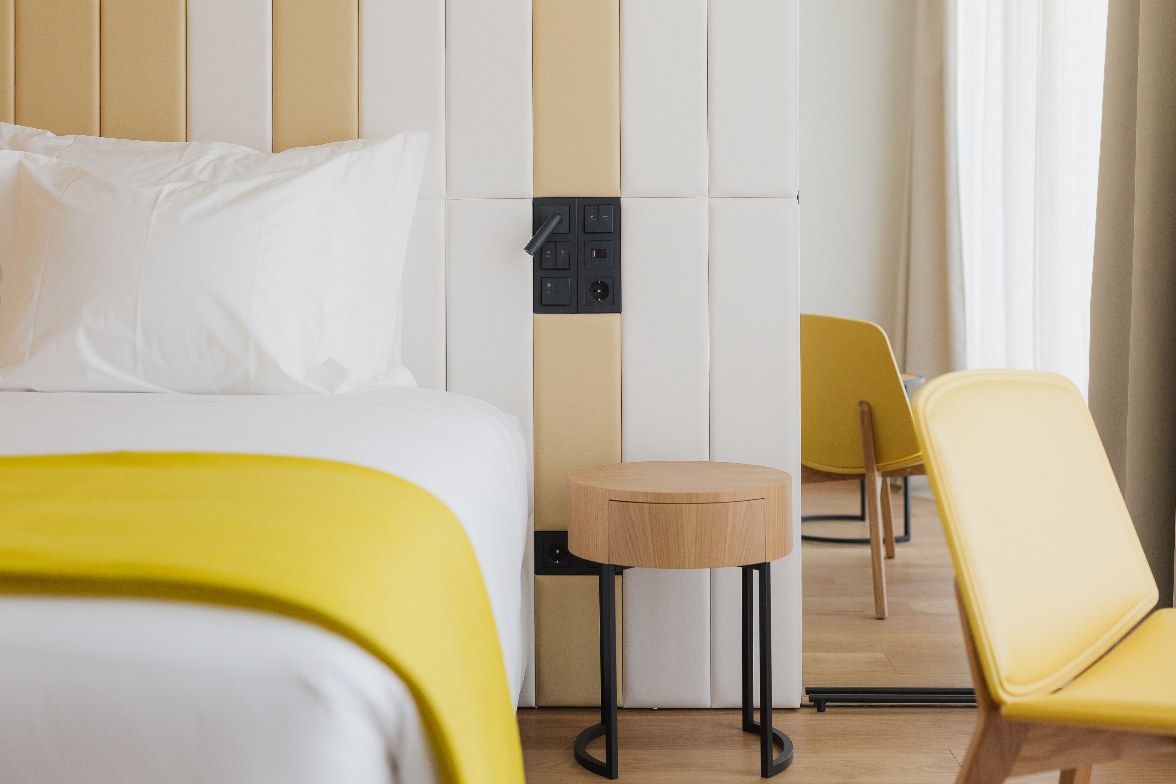 Hotellihuone, jossa on sänky, pieni puinen yöpöytä ja kaksi keltaista tuolia.