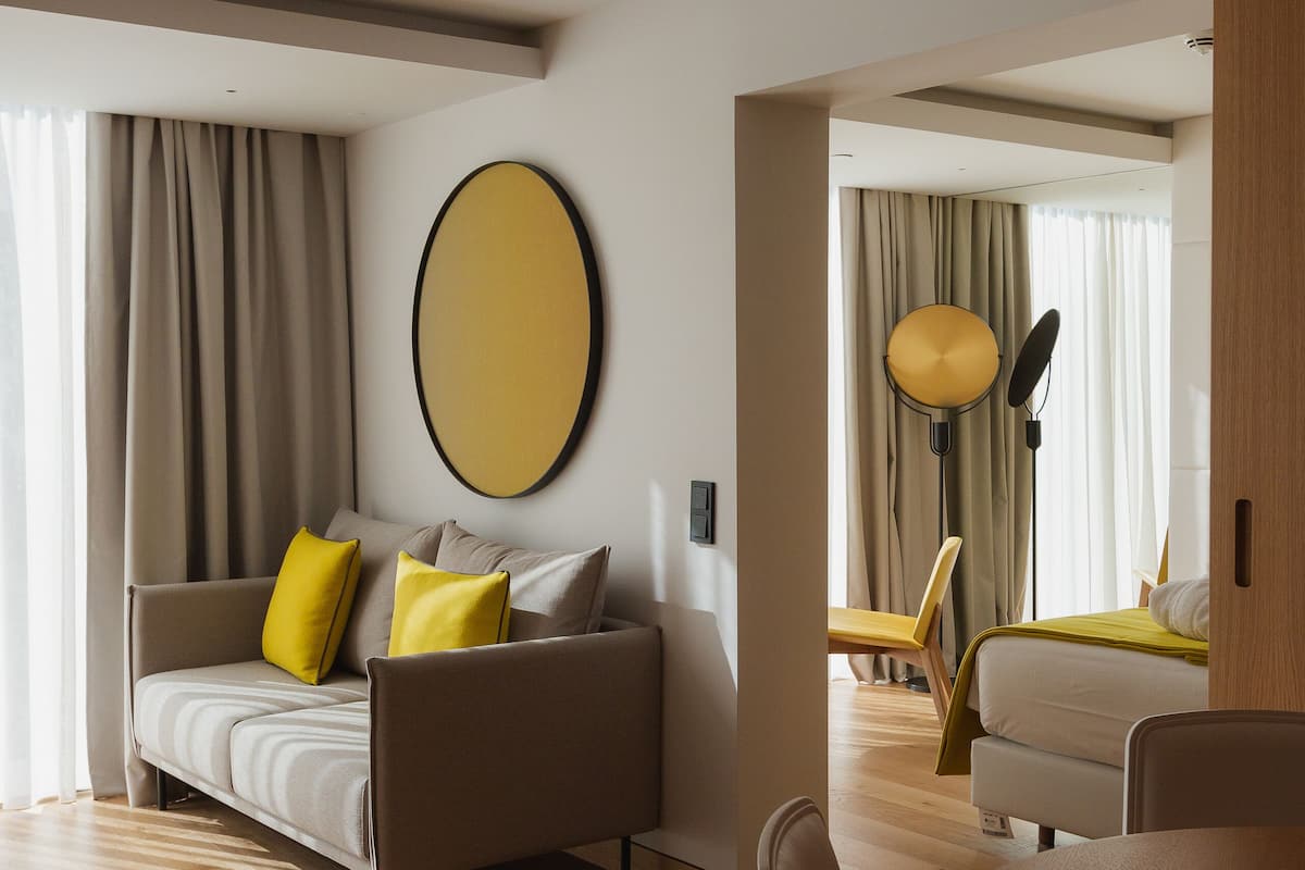 A modern hotel room with a sofa, a round mirror, a dining table, and a large window with curtains.