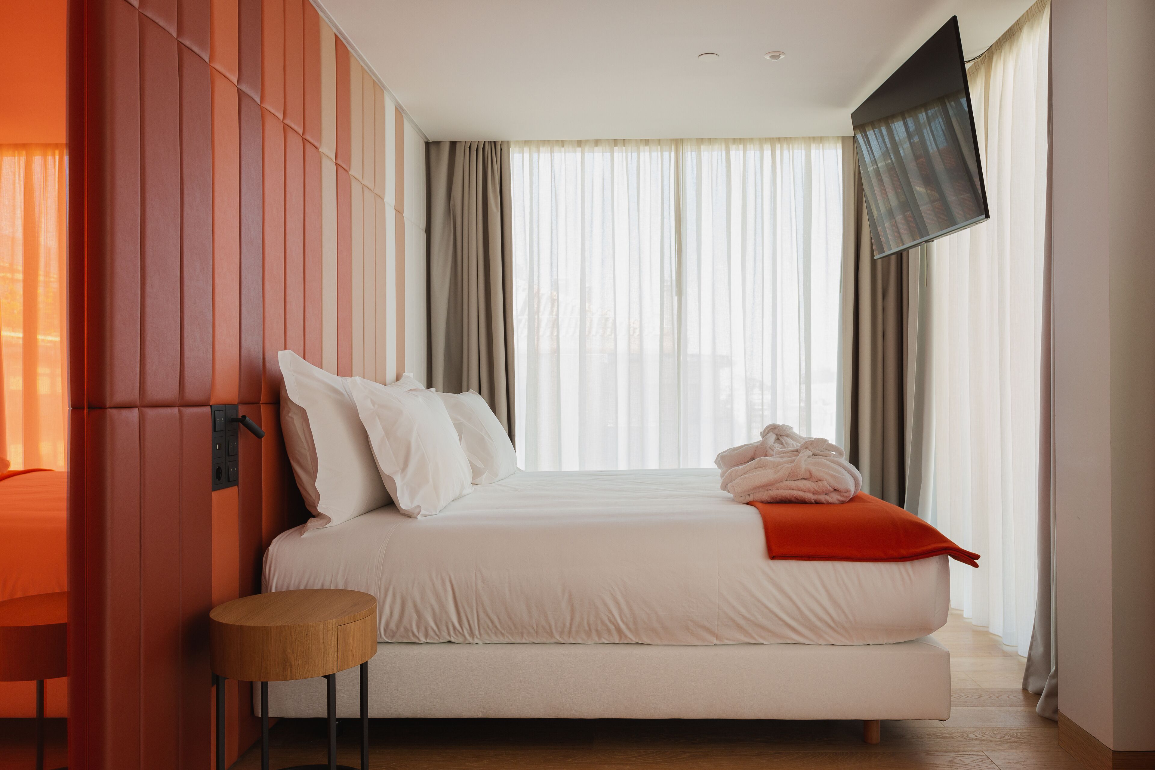 Una habitación de hotel moderna con una cama grande, un televisor de pantalla plana montado en la pared y vistas de la ciudad a través de cortinas translúcidas.
