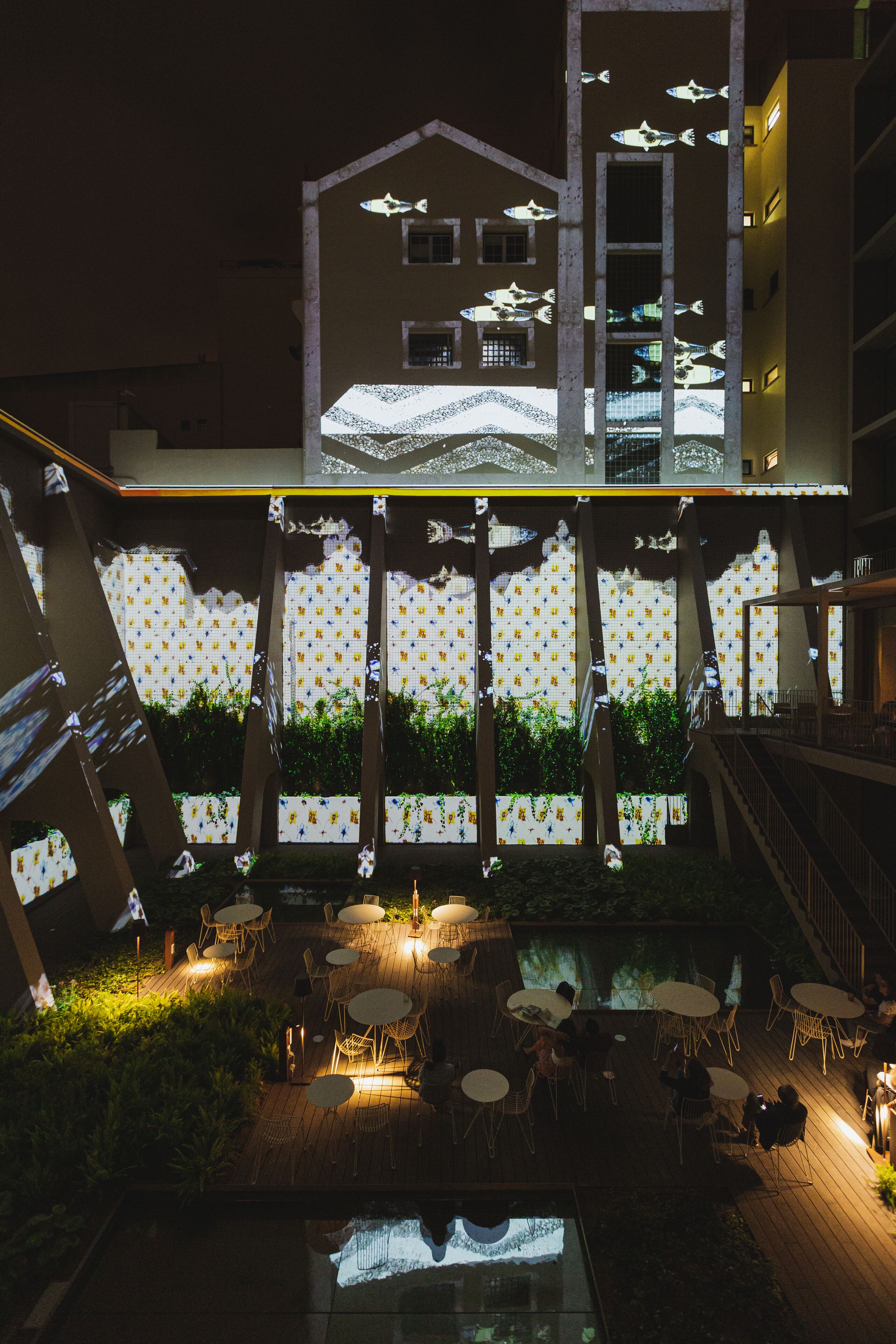 An outdoor patio with tables and chairs, a pool, and a building with fish patterns.