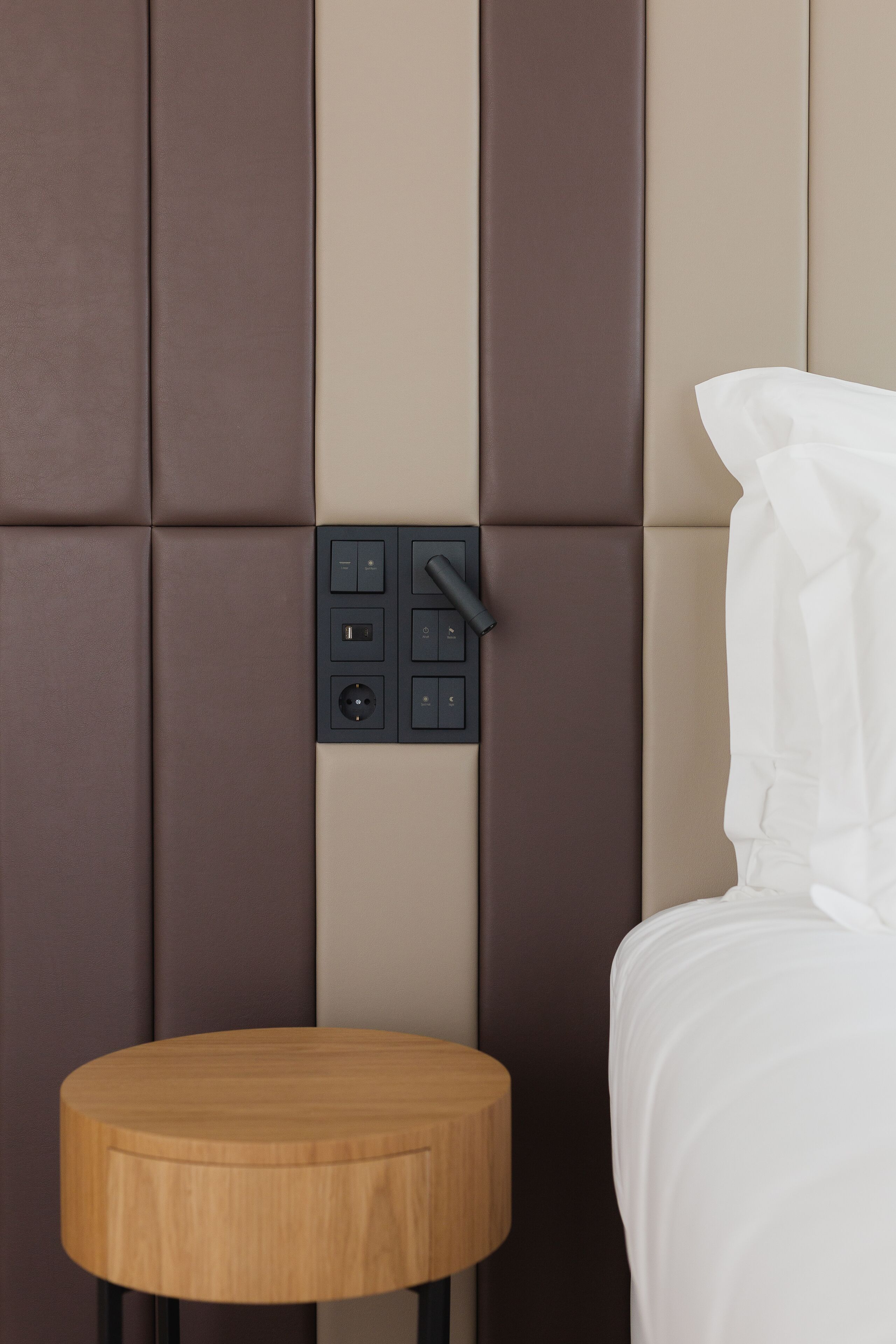 A modern room with a padded wall, a control panel, a wooden side table, and a white bed.