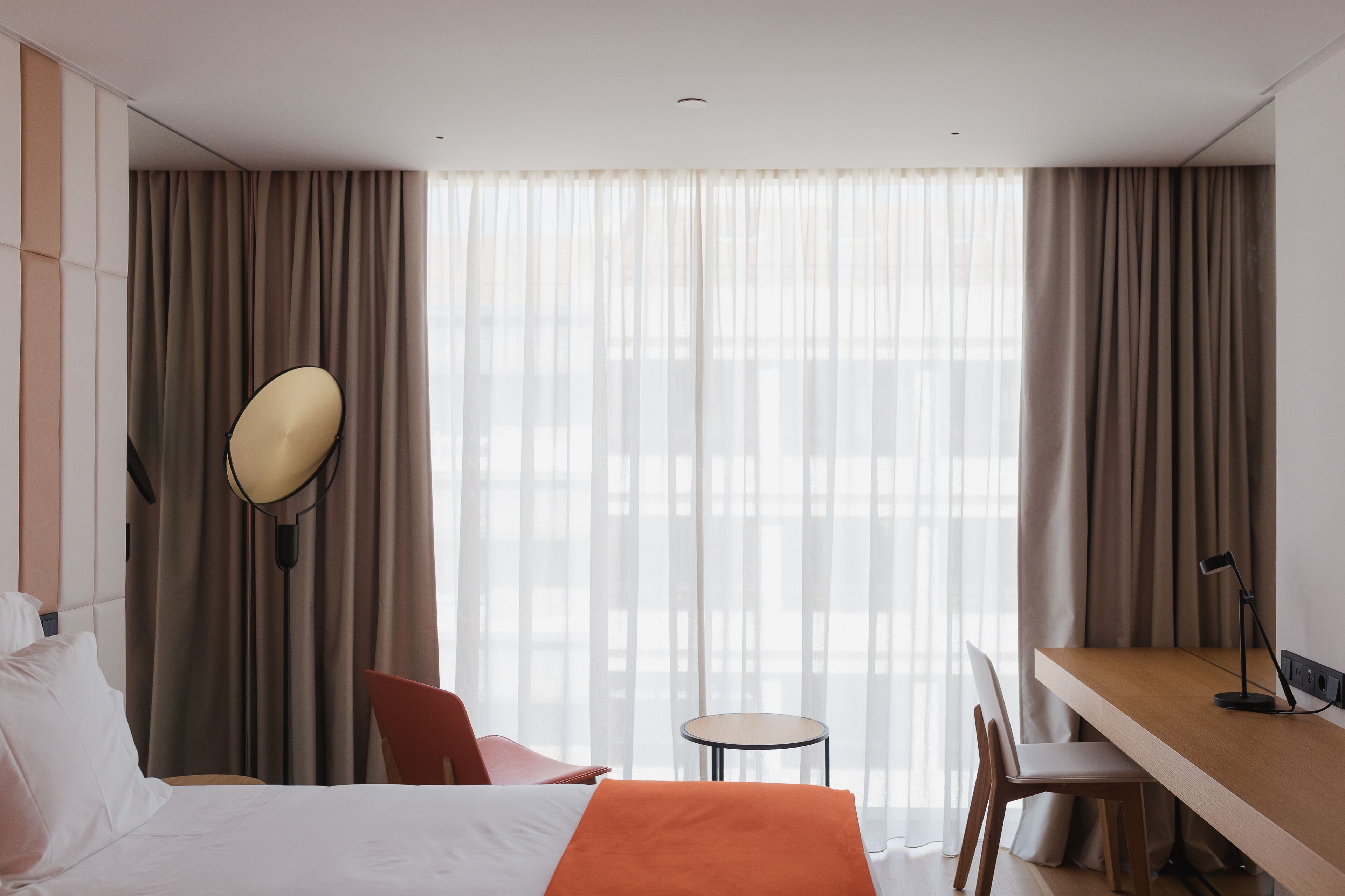 A hotel room with a bed, a desk, a chair, a small table, and a lamp.