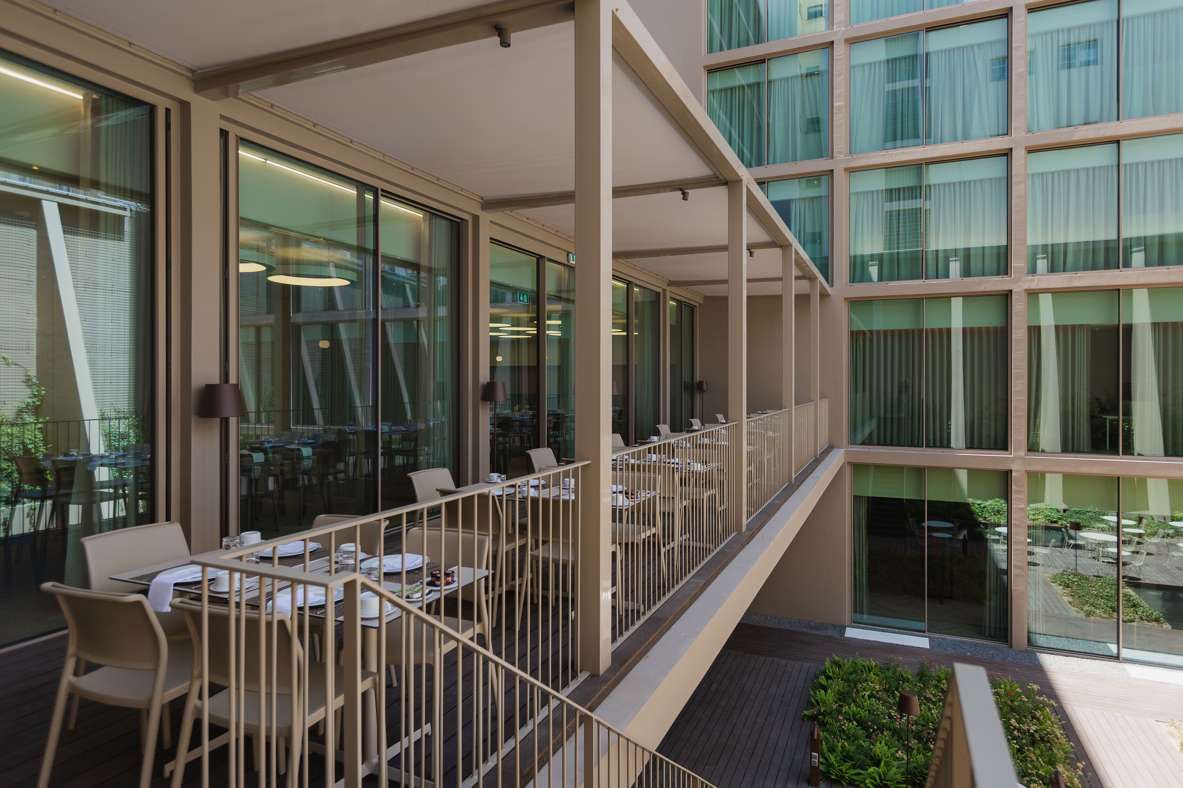 A modern outdoor dining area with tables and chairs, large windows, and a view of the surrounding buildings.